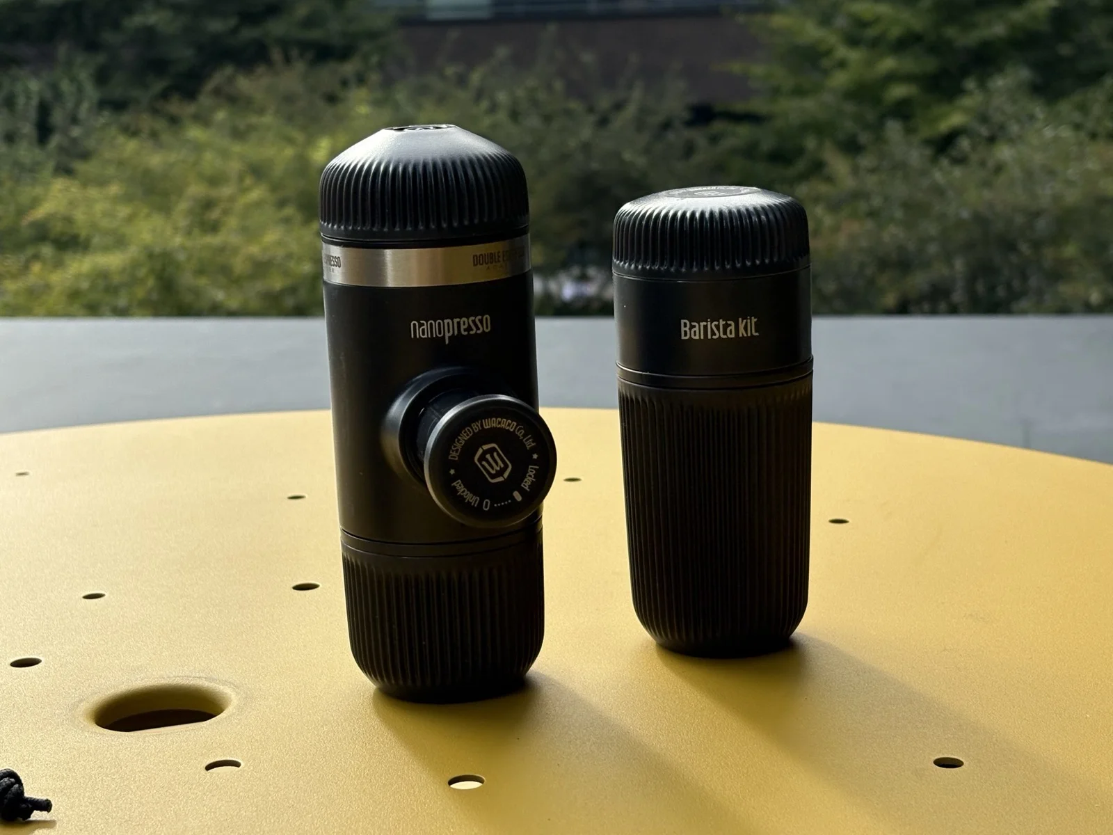 Kávovary Wacaco Nanopresso a Barista Kit vedle sebe na žlutém stolku