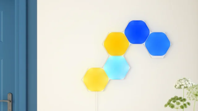 Pět rozsvícených šestiúhelníkových panelů Nanoleaf Shapes ve žluté a modré barvě na světlé stěně nad jídelním stolem